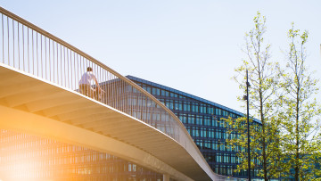 Een fietser fietst over een brug in een stad, met op de achtergrond architecturale hoogbouw en bomen.