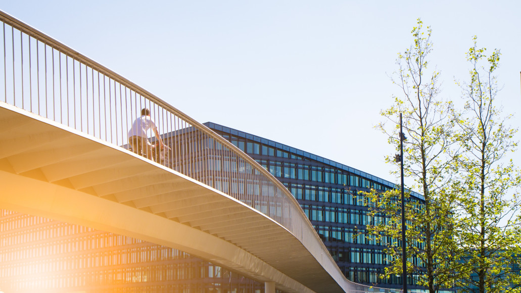 Een fietser fietst over een brug in een stad, met op de achtergrond architecturale hoogbouw en bomen.